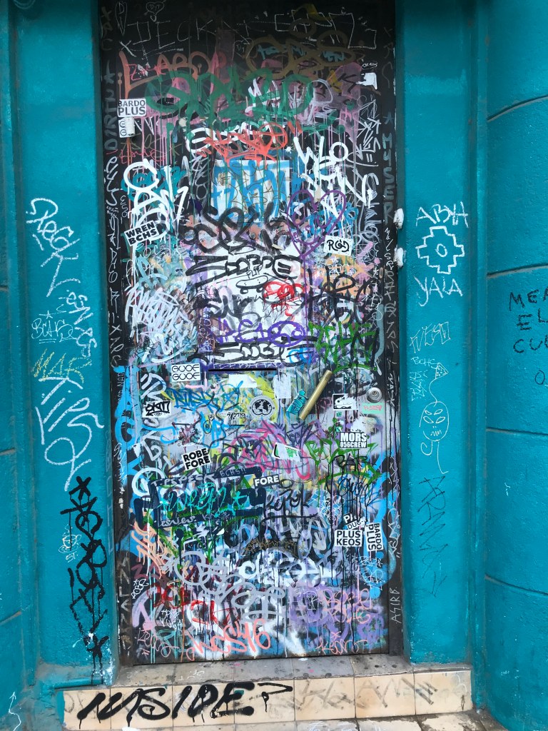 Una puerta negra, que casi no se ve negra porque está repleta de rayados de personas. Se ve parte del marco de cemento del edificio color azul petróleo. Son muchos grafitis que se vuelven ilegibles. Abajo, en la entrada del piso de cerámidas, la parte frontal del piso cerámico color cr5ema se lee el grafitti "INSIDE¨. También destaca uno que está al lado derecho que dice Abya Yala, con un símbolo como andino (no sé de dónde es, perdón)